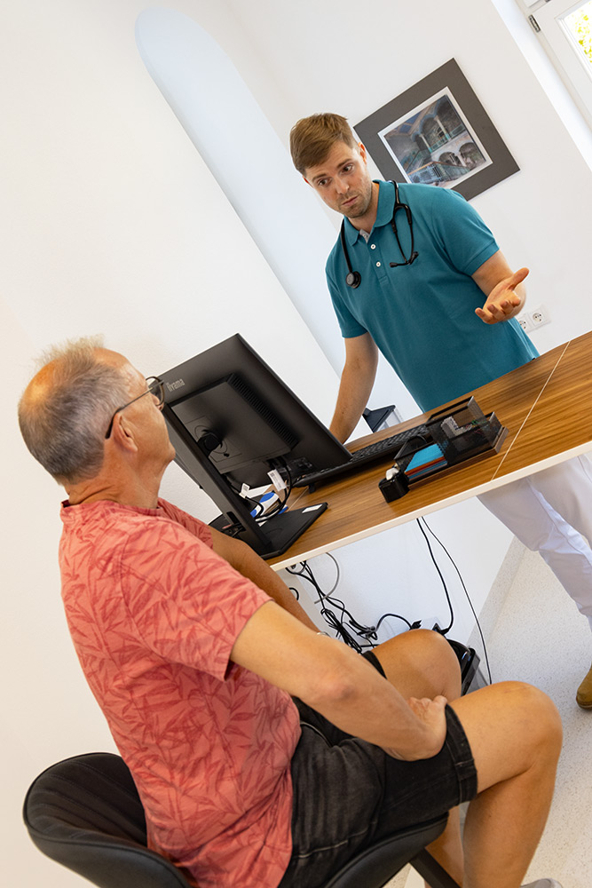 Dr. David Wittmann stehend hinter dem Schreibtisch berät einen Patienten, der davor sitzt.